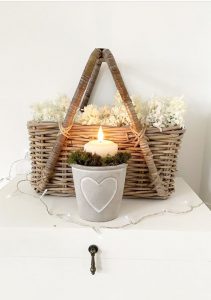 Festive Christmas candle decorations. Luxury LED pillar candle inside a plantpot surrounded by moss with a basket of foliage in the background